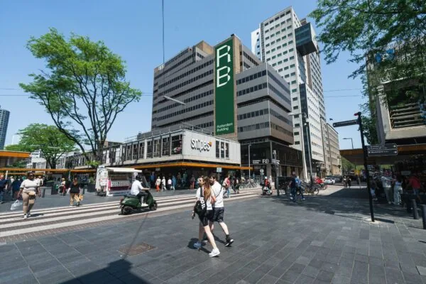 Drukke winkelstraat bij de Aert van Nesstraat in Rotterdam met voetgangers, fietsers en het Rotterdam Building op de achtergrond.